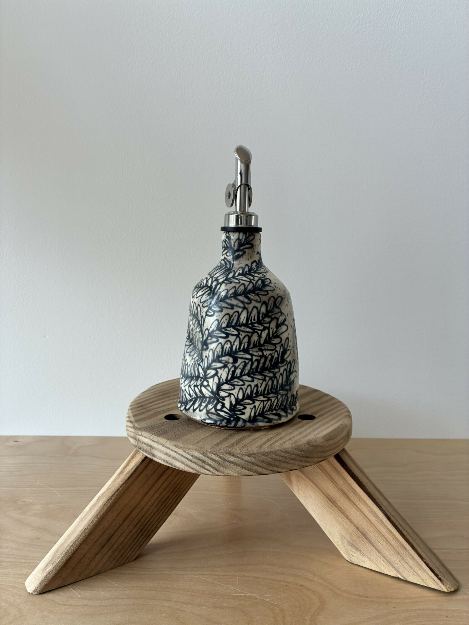 Ceramic bottle with black and white leaf pattern on a wooden stool, against a plain white background.
