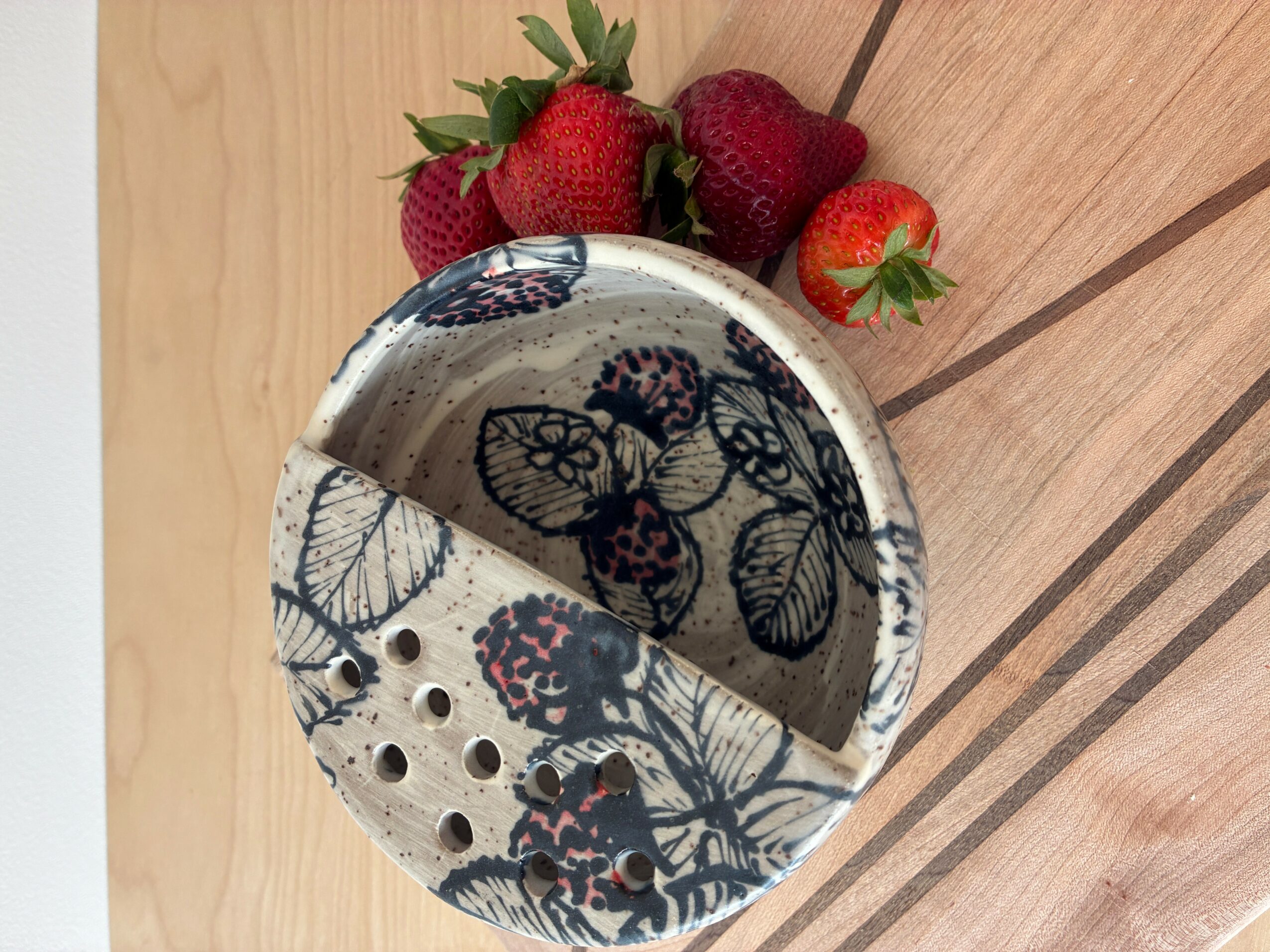 A round container with a butterfly and leaf pattern, filled with strawberries, on a wooden surface.