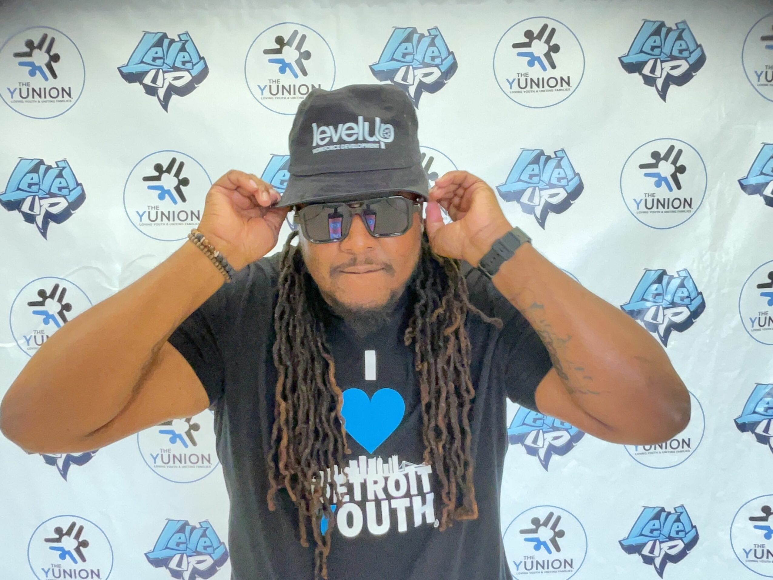 Person with long dreadlocks wearing glasses and a black shirt, holding a camera, upside down, in front of a patterned background.