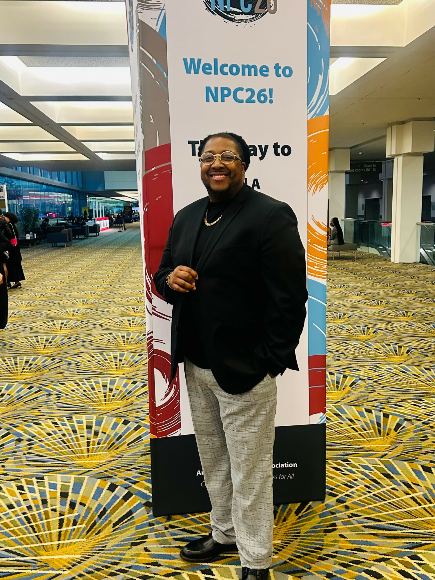 Smiling man in black blazer and light pants standing in front of a colorful NPC26 sign at a conference center.