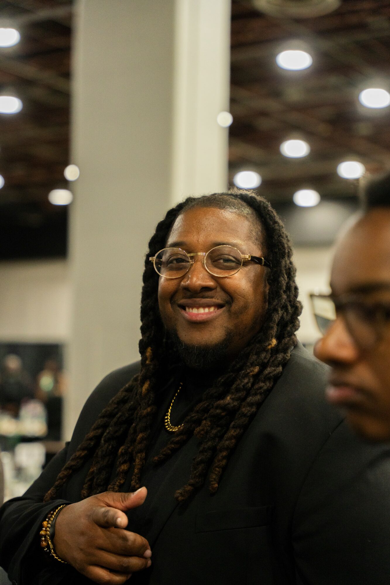 Smiling person with glasses and long dreadlocks, wearing a black outfit, in an indoor setting with bright lights.