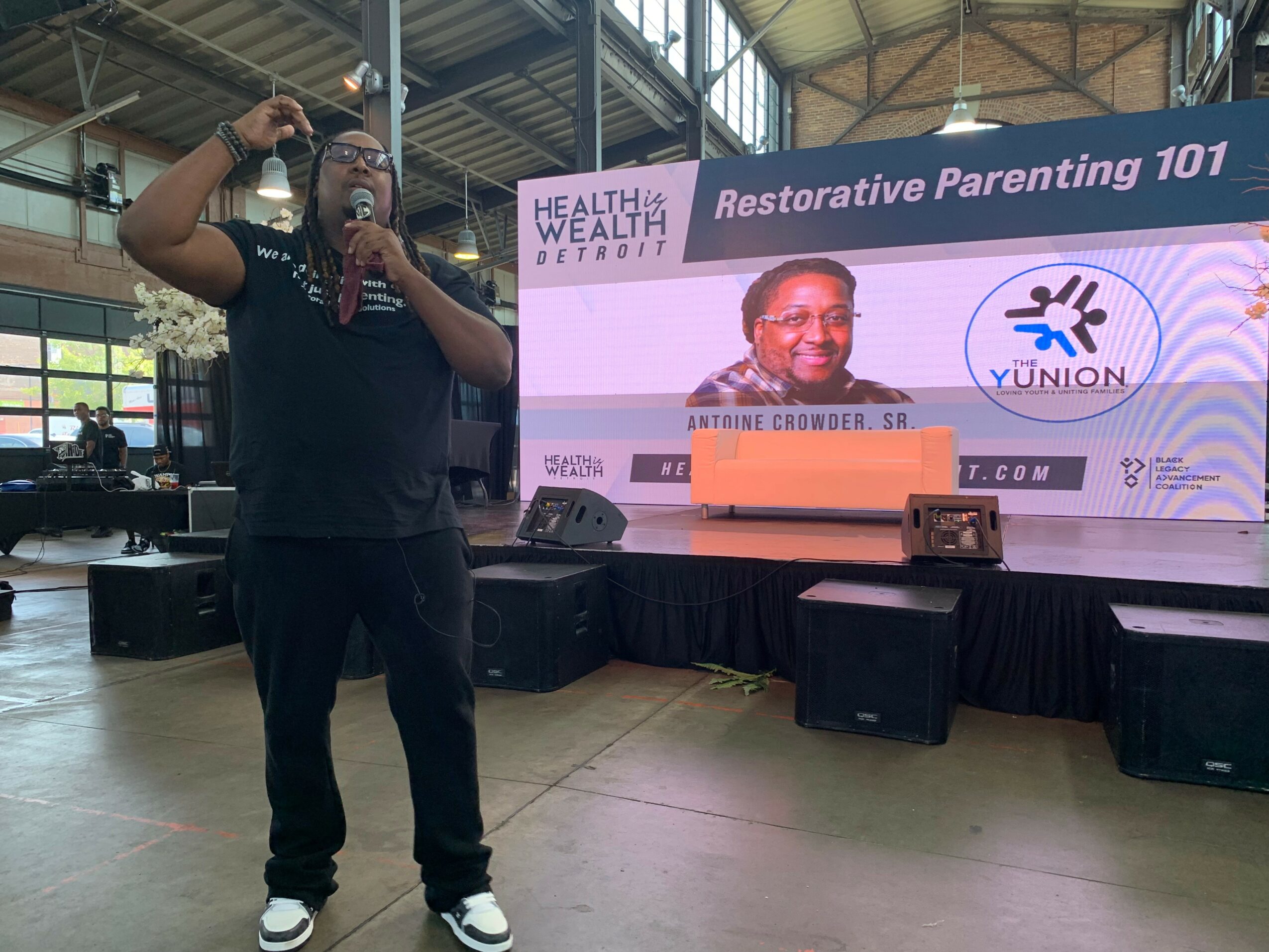 Man speaking on stage with a large screen behind him displaying a presentation titled 'Restorative Parenting 101' and a logo.