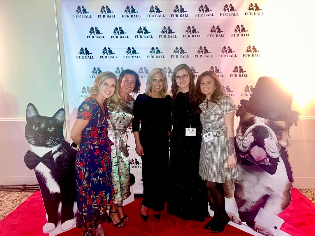 Five women stand together on a red carpet in front of a backdrop with logos, smiling for the photo.