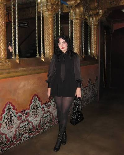 Woman with dark hair in black outfit and patterned tights standing indoors near ornate decor.