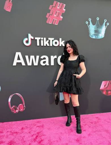 Woman in black dress and knee-high boots standing on pink carpet at TikTok Awards event, with decorative icons on dark background.