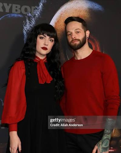 A woman with dark hair and red lipstick and a man with a beard and tattooed arm stand together, both wearing red and black clothing.