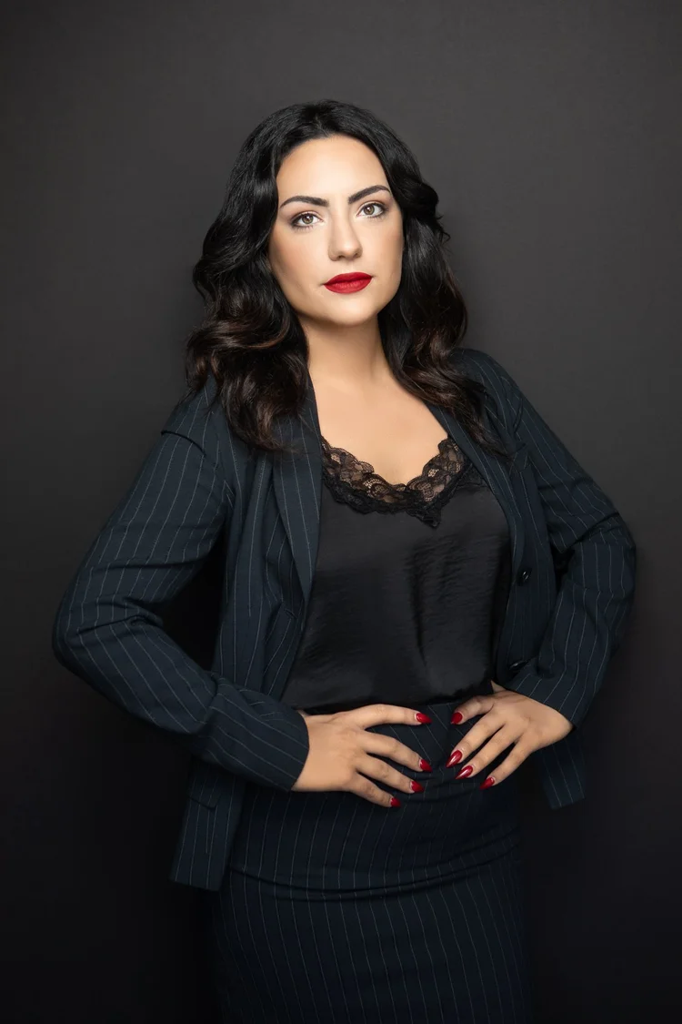 Woman with dark wavy hair, red lipstick, wearing a black top with lace, and a dark blazer, standing against a dark background.