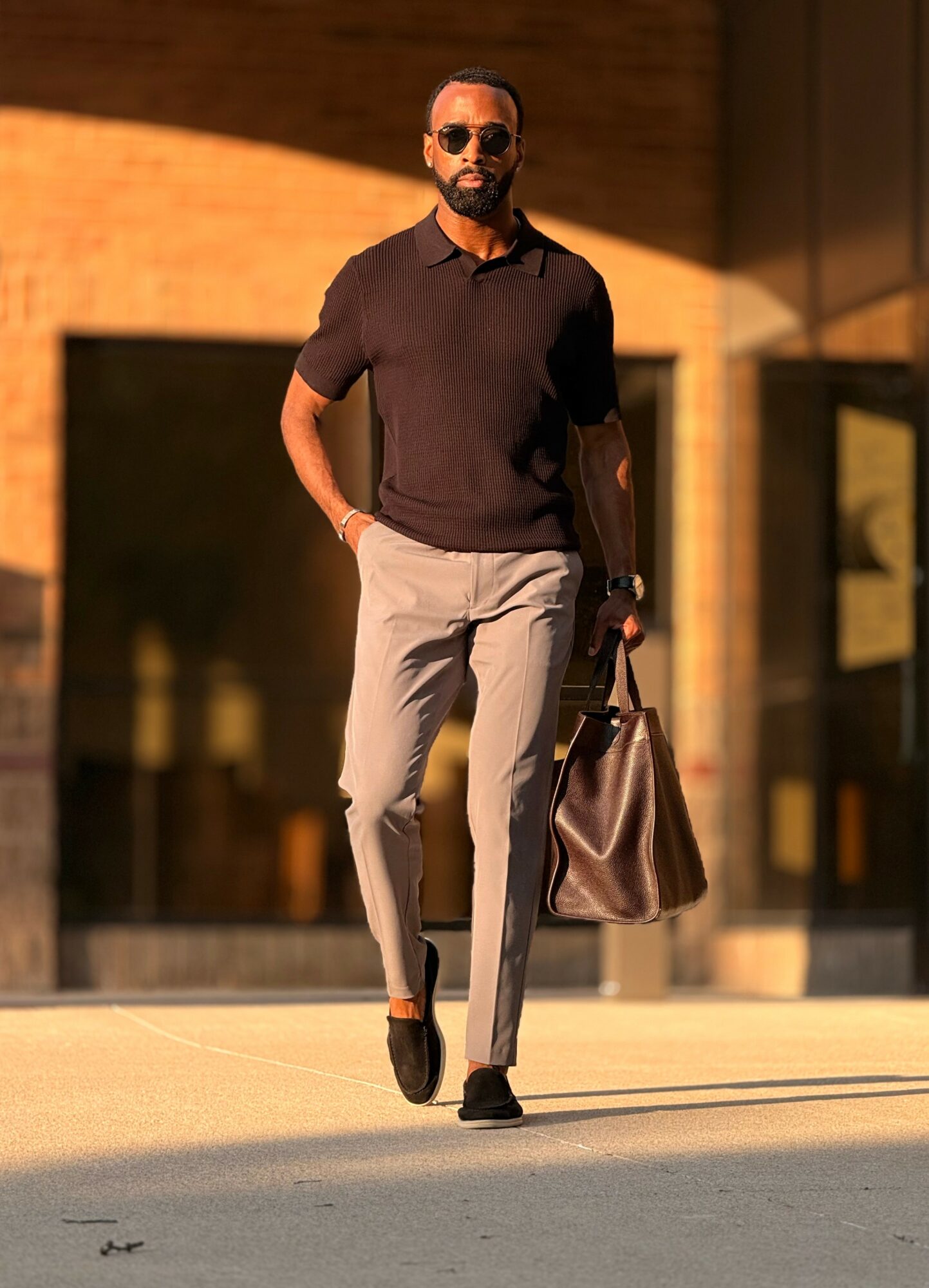 Man walking outdoors in sunlight, wearing sunglasses, black shirt, gray pants, carrying a large brown bag.