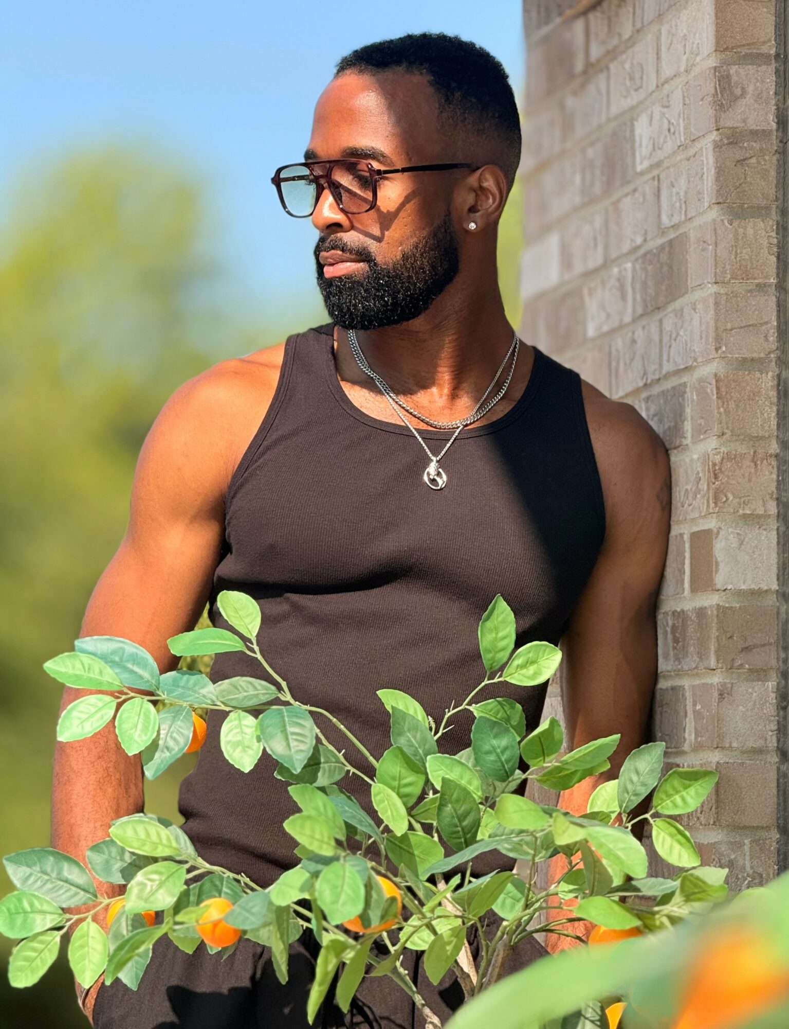 Man with glasses and a beard leaning against a brick wall outdoors, wearing a black sleeveless shirt, surrounded by green leaves.