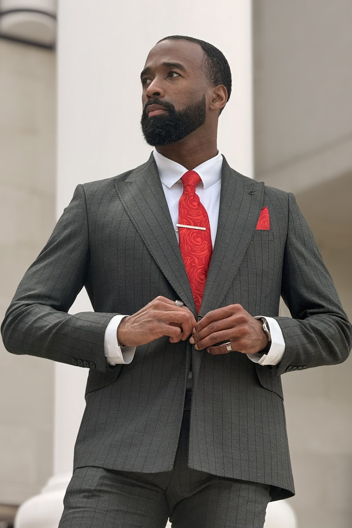Man in a gray suit with a red tie and pocket square, standing outdoors with a neutral background.