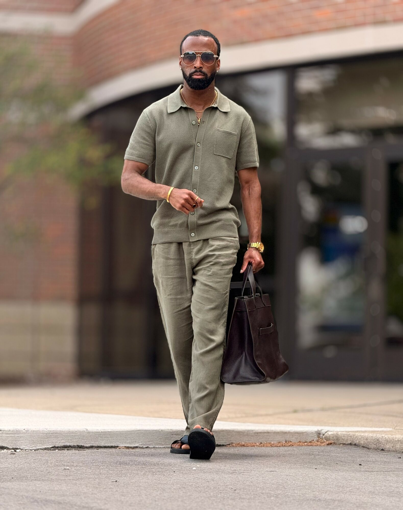 Man walking on sidewalk wearing sunglasses, olive green outfit, carrying a dark bag, in front of a building with glass windows.