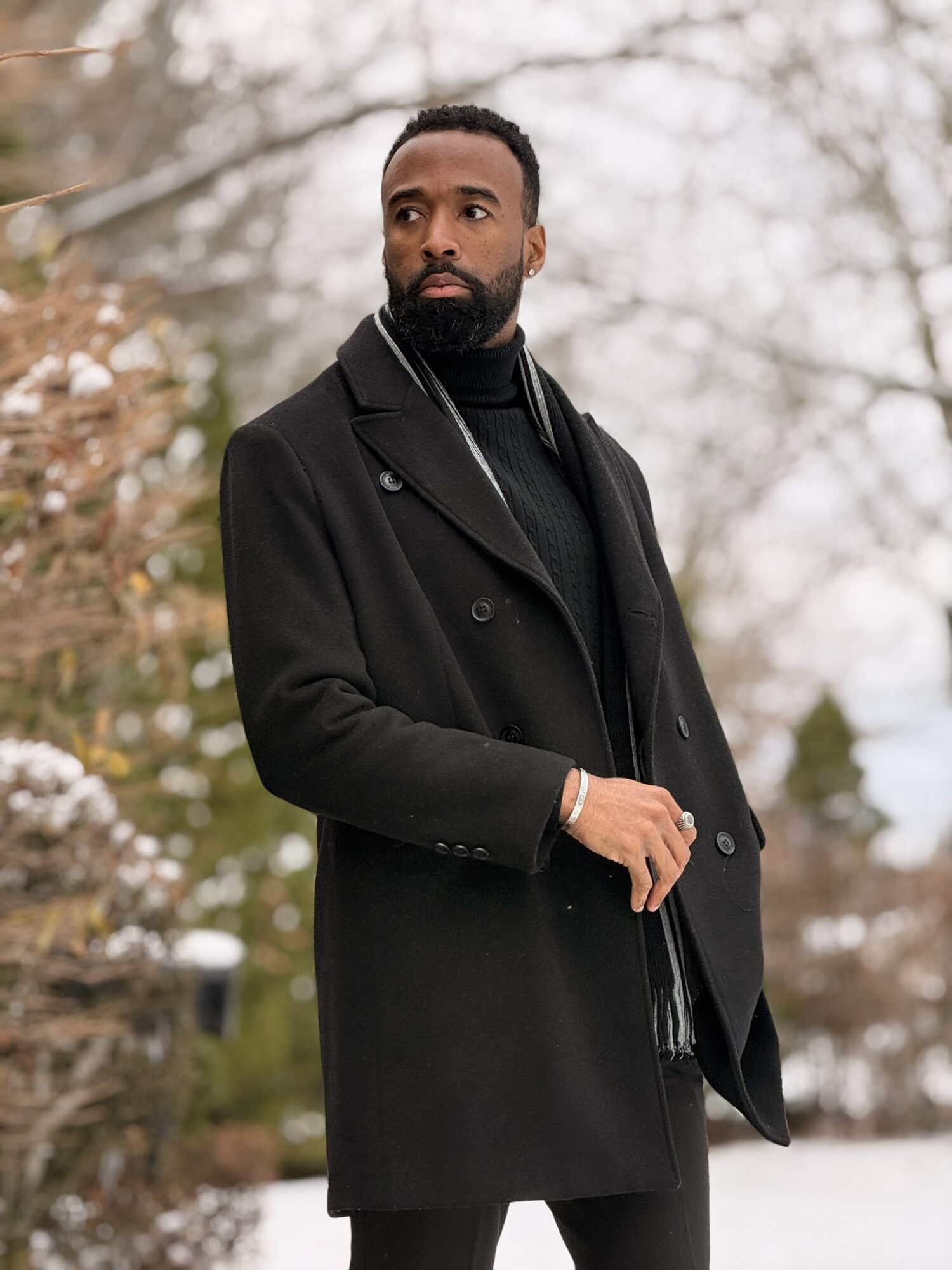 Man with a beard wearing a black coat and scarf outdoors in a winter setting.