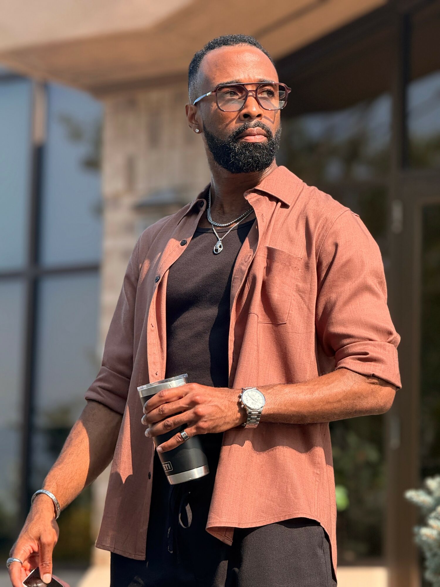 Man with glasses and a beard holding a drink, standing outdoors near a building, wearing a brown shirt over a black top.