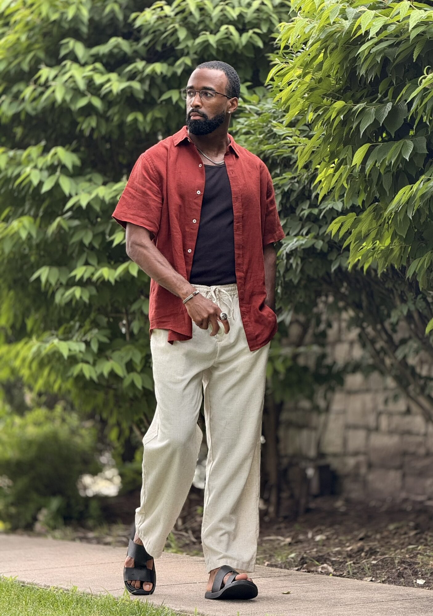 Man walking outdoors with green trees, wearing a red shirt, black t-shirt, beige pants, and sandals.