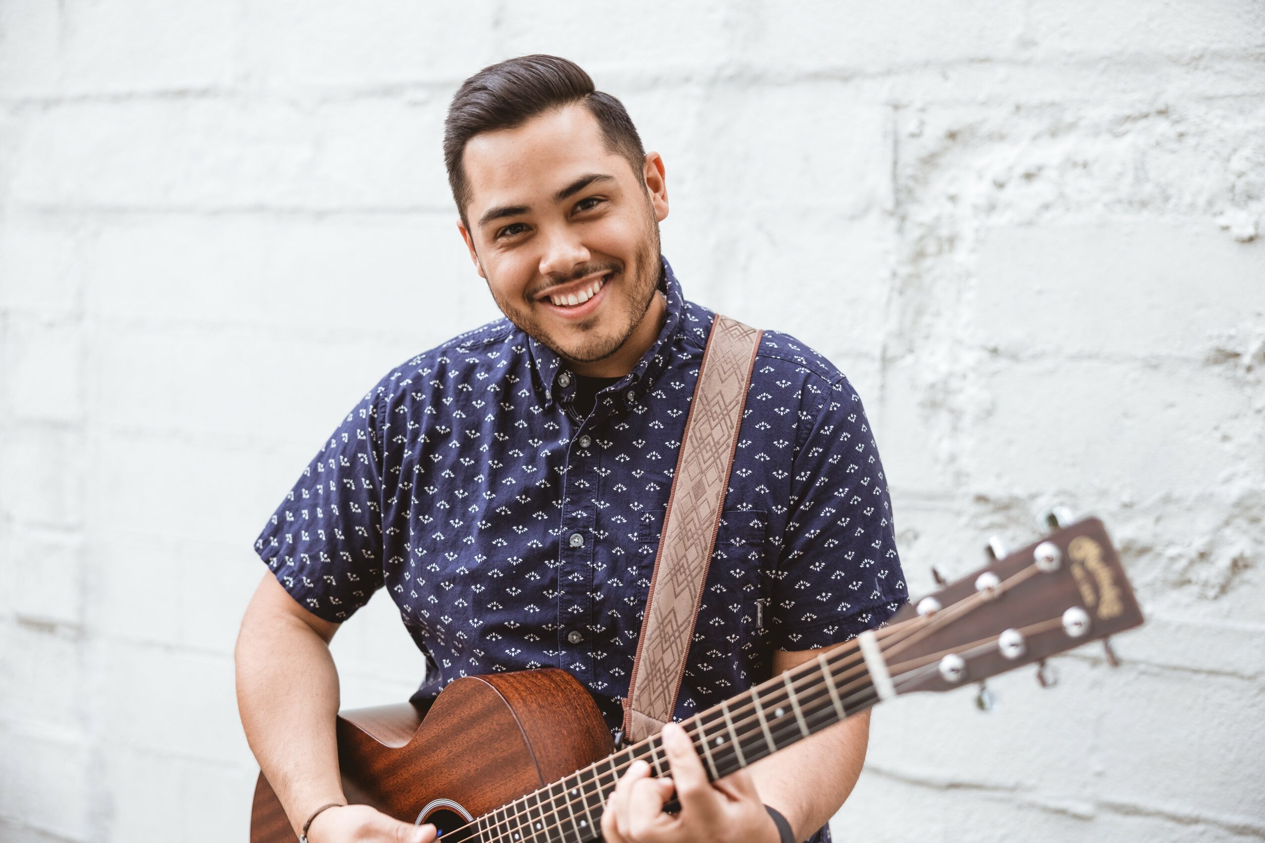 Young man smiling, holding an acoustic guitar, wearing a dark patterned shirt, standing against a white brick wall.