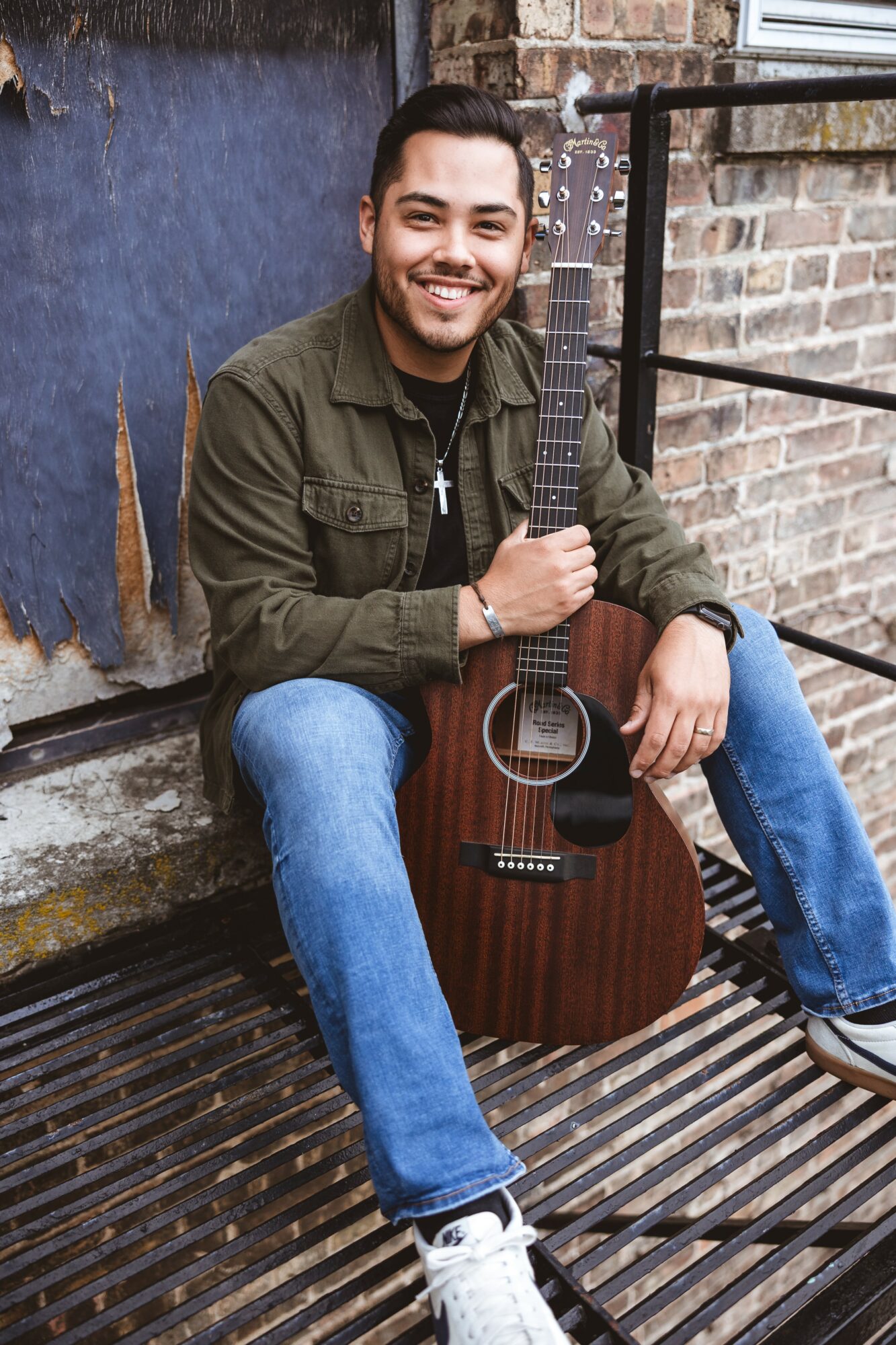 Young man sitting outdoors with an acoustic guitar, smiling, against a brick wall and metal railing.