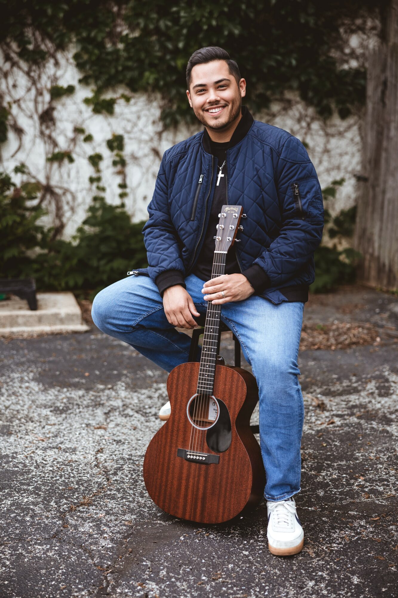 Man smiling outdoors holding an acoustic guitar, standing on pavement with trees in background.