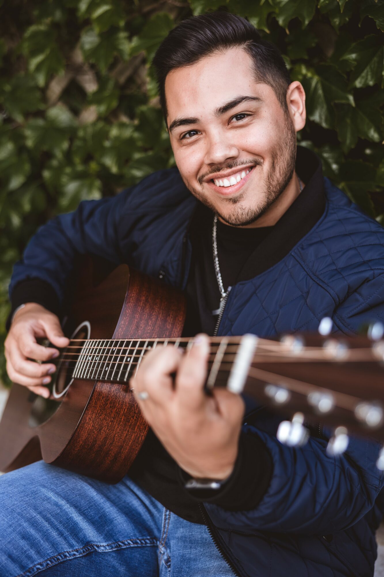 Young man smiling while playing an acoustic guitar outdoors, surrounded by green foliage.