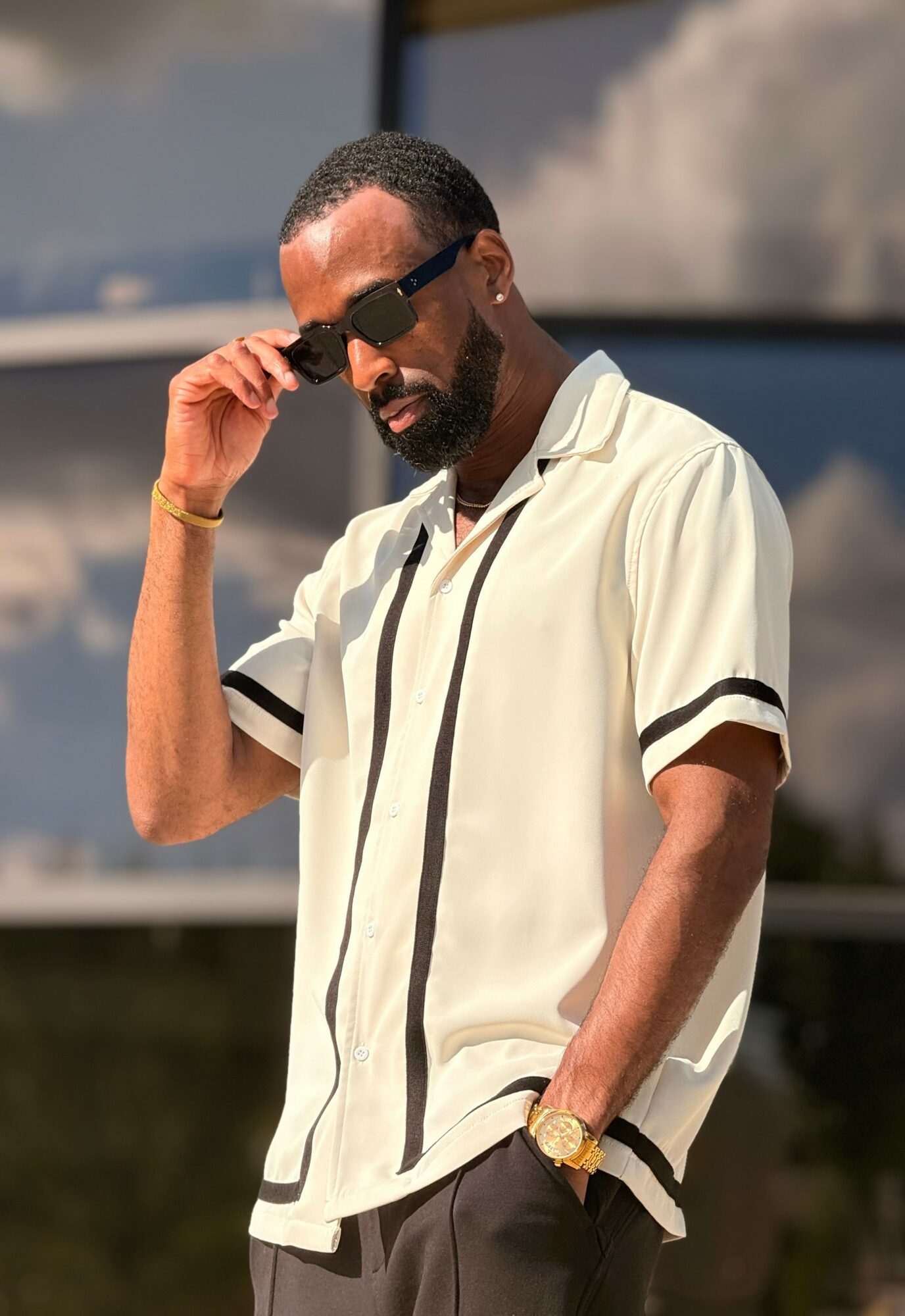 Man with sunglasses adjusting his glasses outdoors, wearing a light-colored short-sleeve shirt and a gold watch.