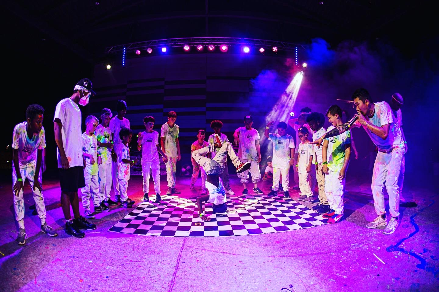 Group of people dancing on a checkered floor under colorful stage lights, with smoke effects in the background.