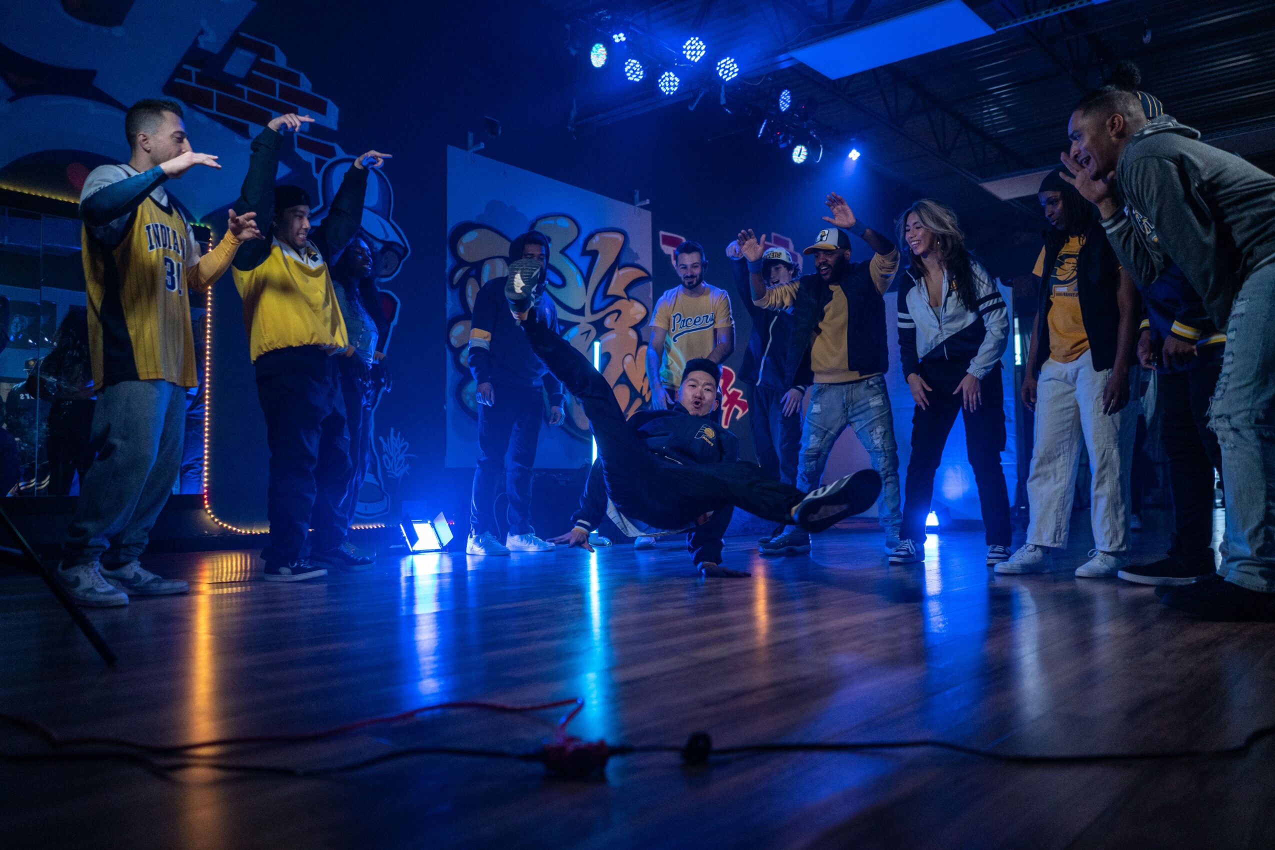Group of people on stage, some standing and one person lying on the floor, with blue stage lighting and a dark background.