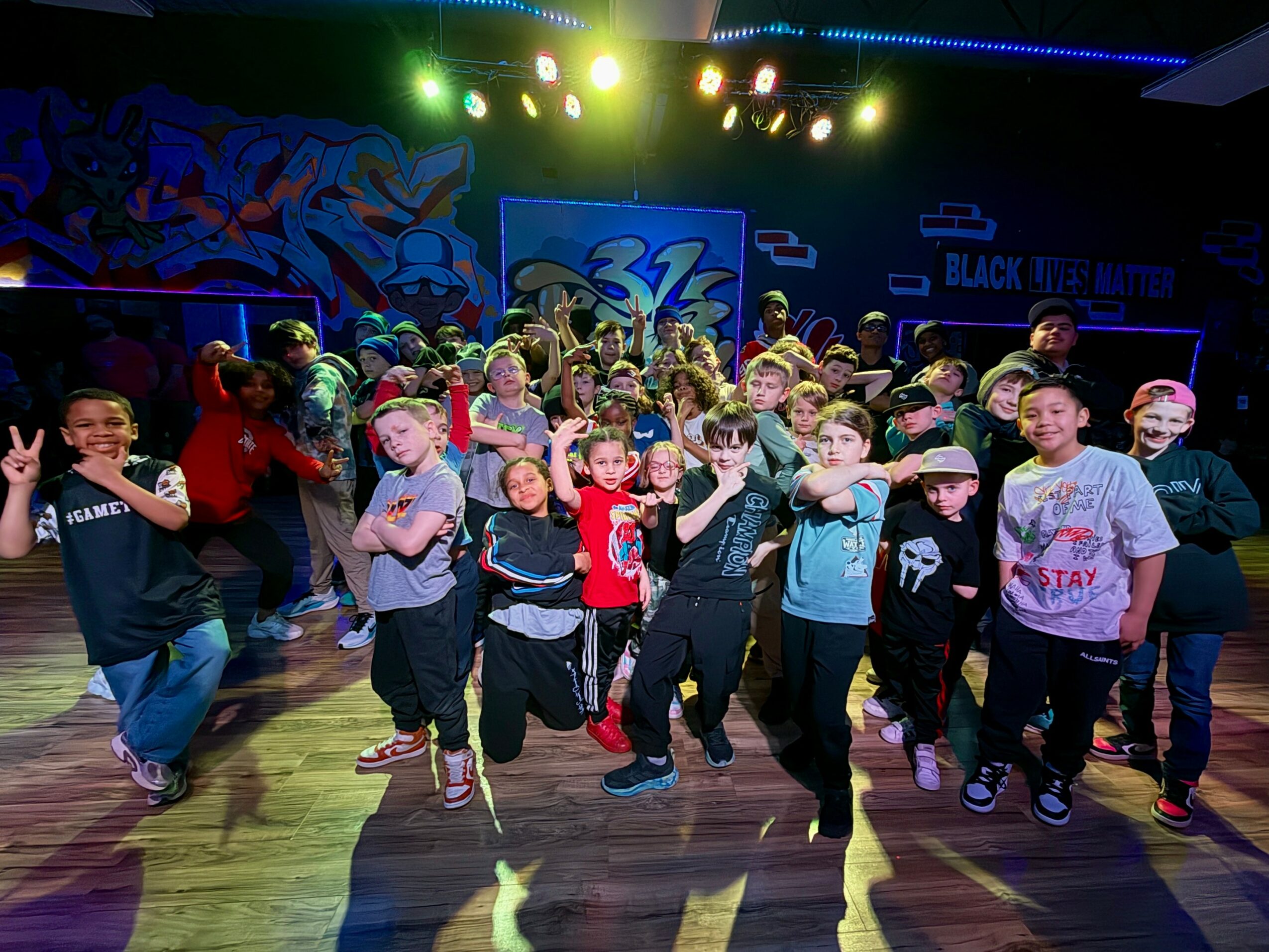 Group of children dancing in a room with colorful graffiti and stage lights, some wearing hats and casual clothes.