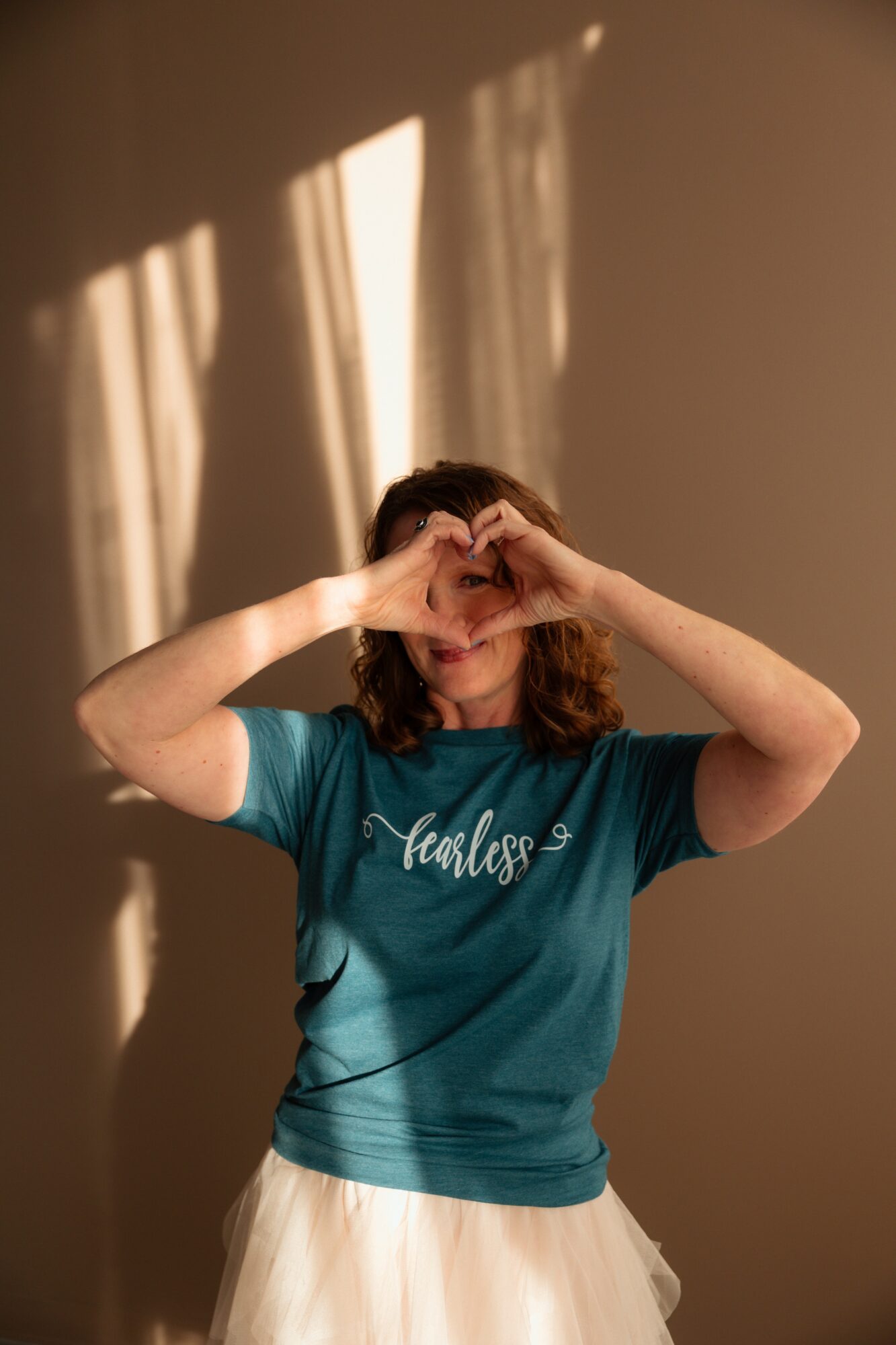 Person with curly hair making a heart shape with hands over face, wearing a teal shirt and white skirt, standing against a wall with shadows.