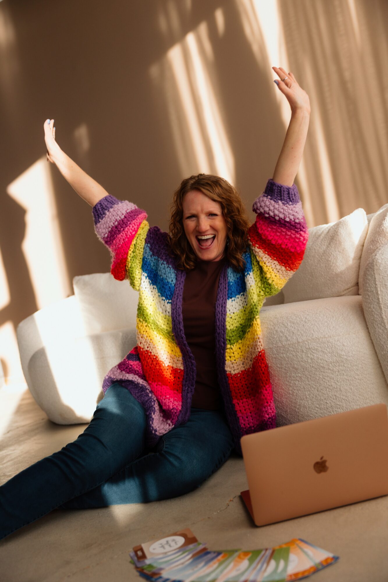 Woman sitting on the floor with arms raised, smiling, in a room with sunlight and curtains, laptop nearby.