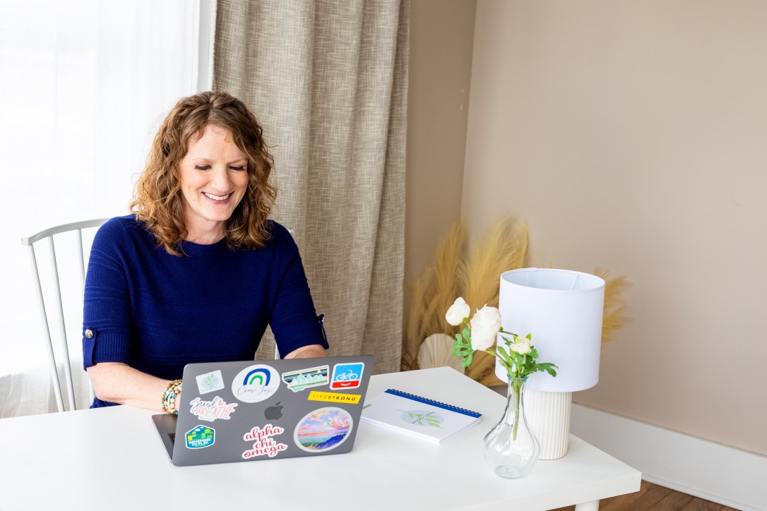 Woman with curly hair smiling at a laptop on a white desk, in a room with beige walls and curtains.