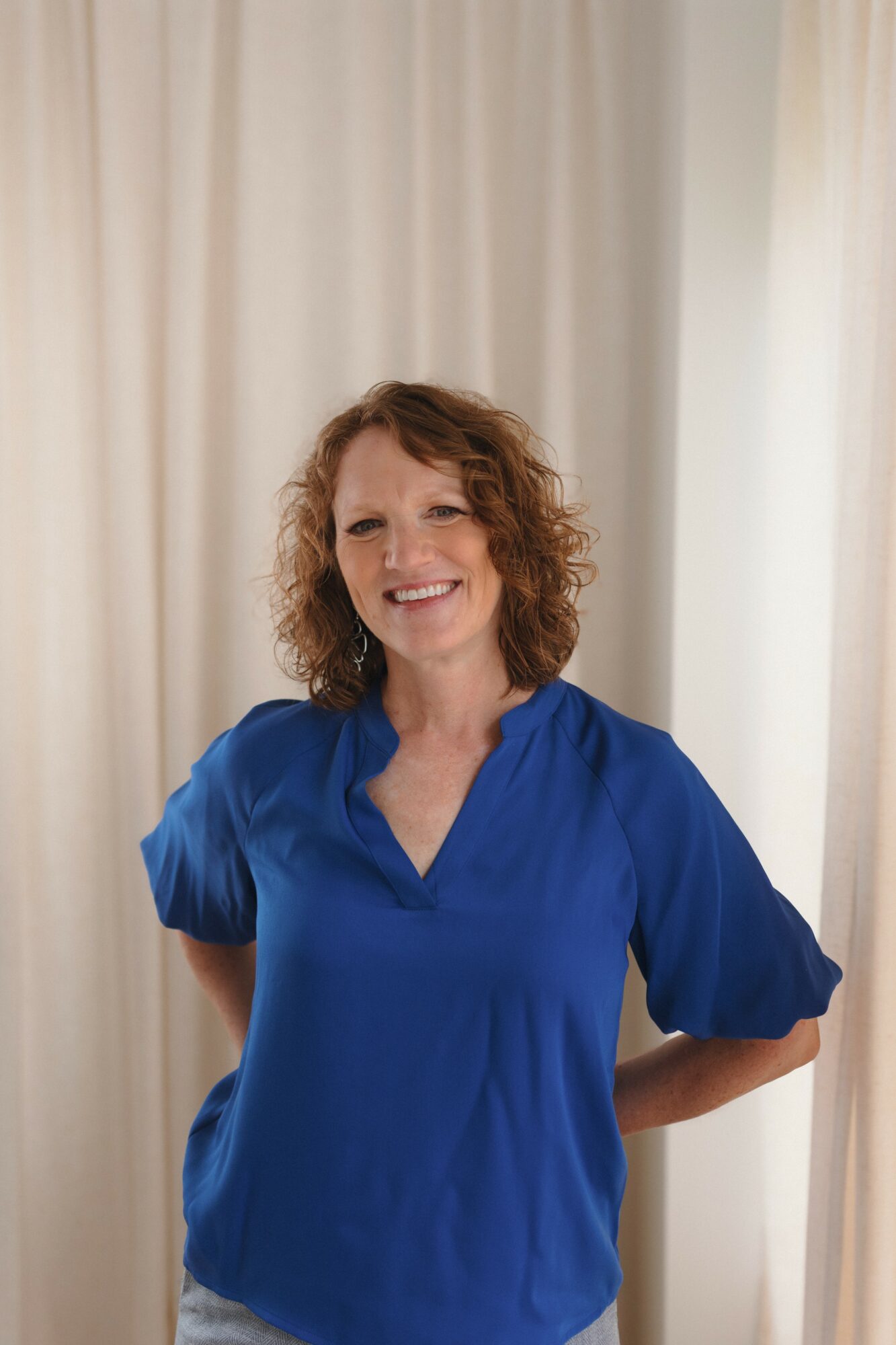 Woman with curly hair smiling, wearing a blue top, standing in front of light-colored curtains.