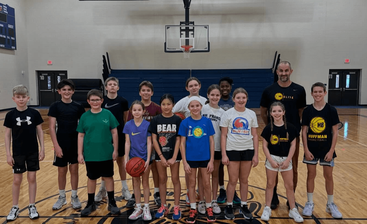 Group of children and coaches in a gymnasium, some holding a basketball, standing in front of a basketball hoop.