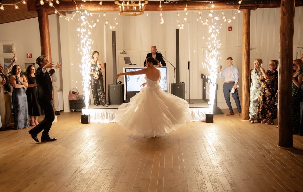 Bride dancing with sparks and guests watching in a decorated hall, DJ in background, wooden floor, festive atmosphere.