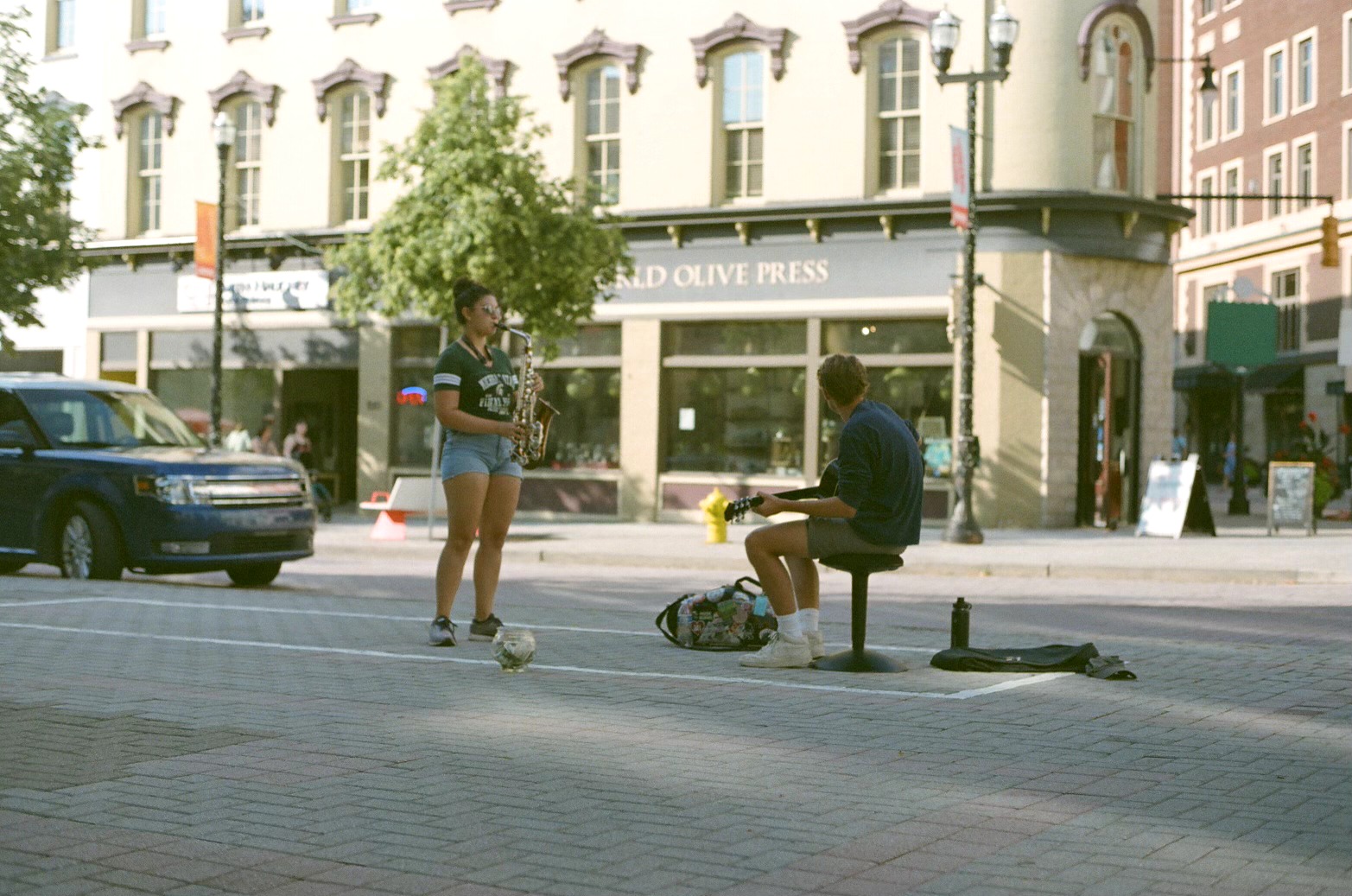 Two people on a city sidewalk, one standing and one sitting on a bench, with buildings and cars in the background.