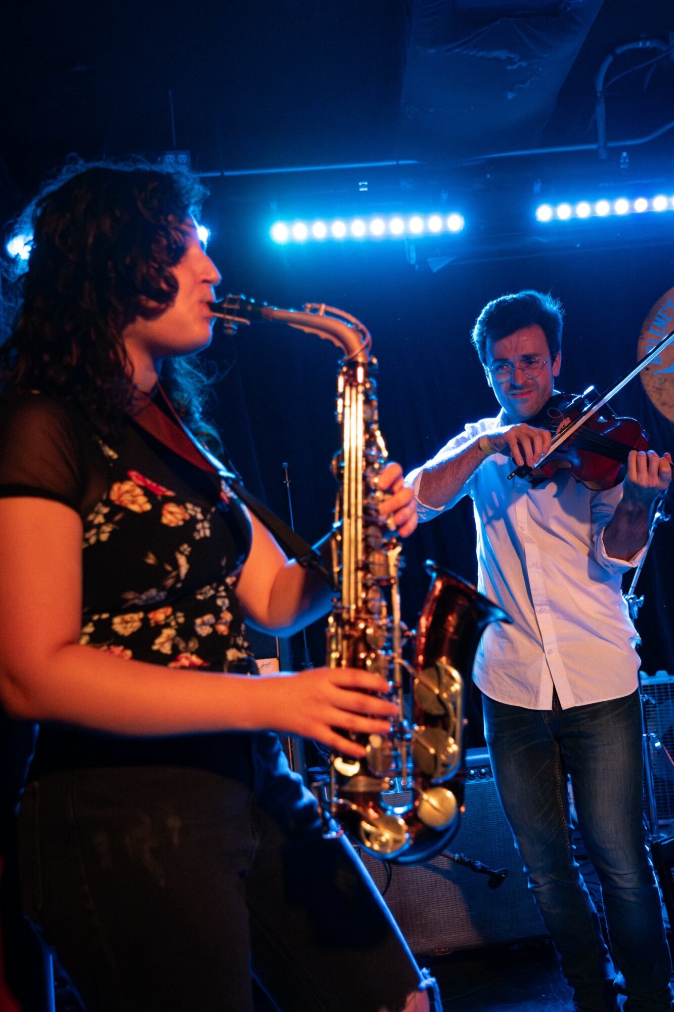 Two musicians perform on stage, one playing saxophone and the other playing violin, with blue stage lighting.