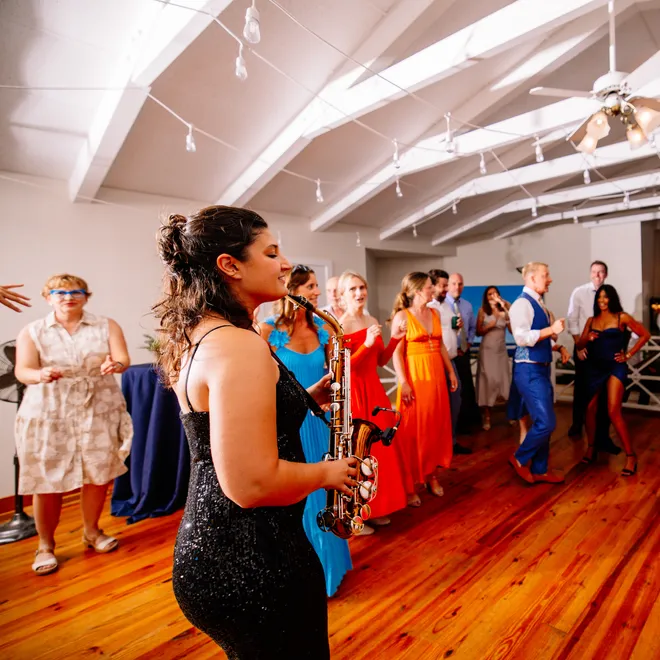 Woman playing saxophone at a dance event with people dancing in the background, wooden floor, ceiling with white beams and string lights.
