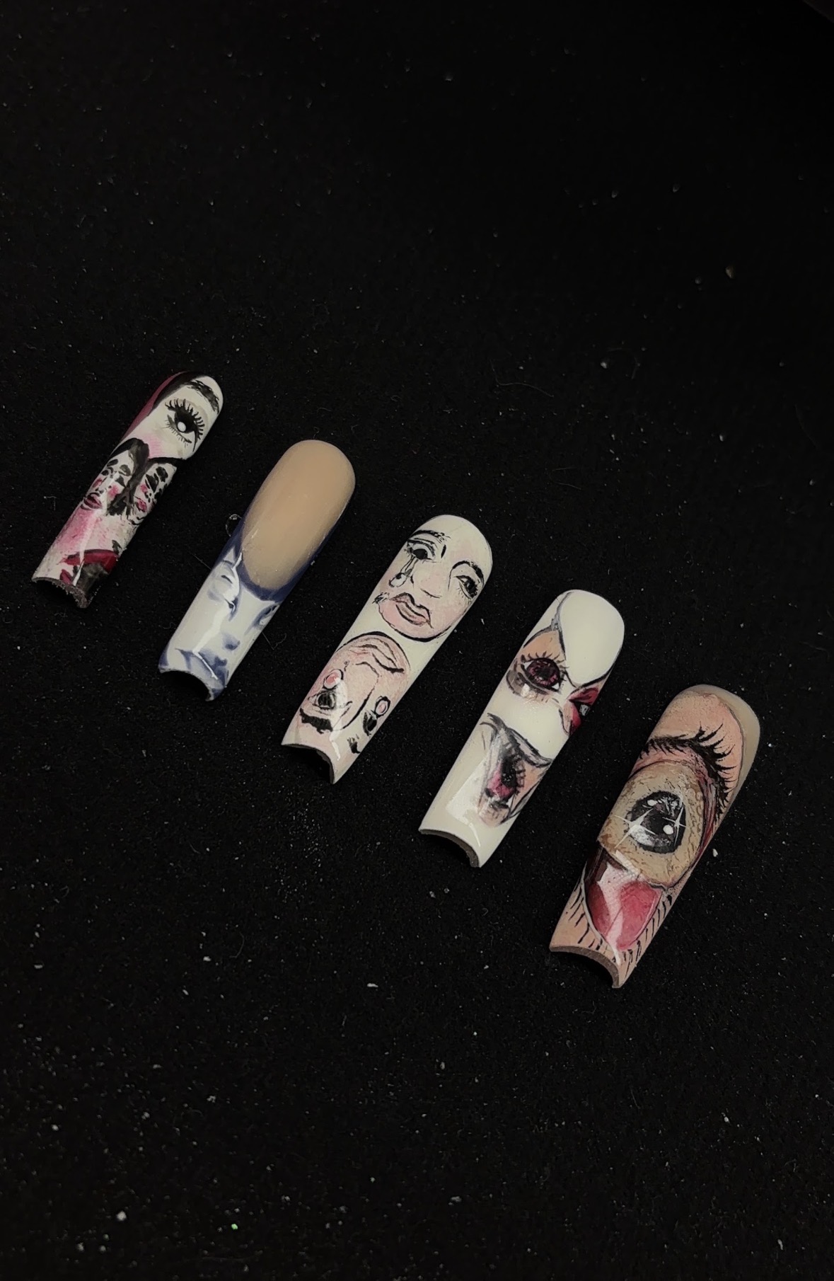 Six false nails with illustrated faces and eye designs, arranged in a row on a black background.