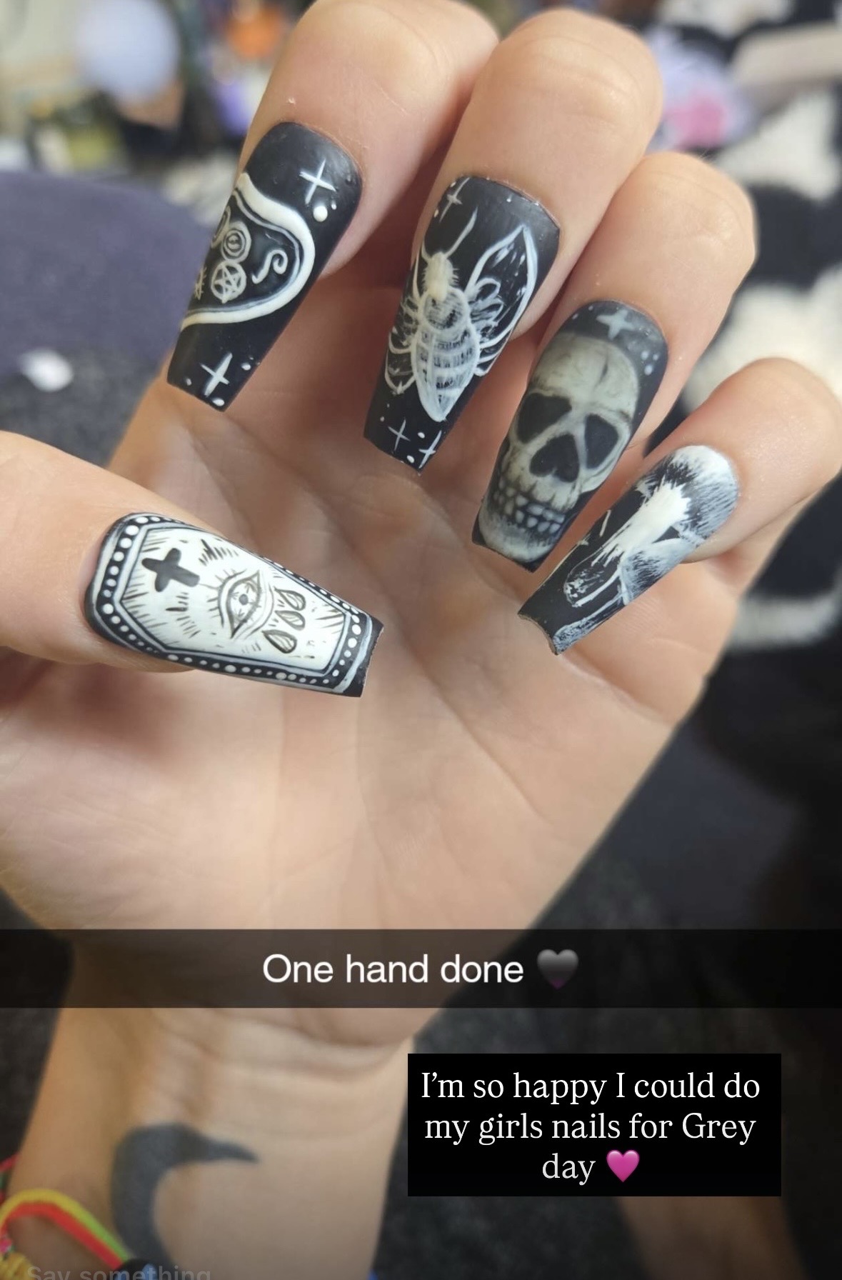 Hand with black and white tattoo-style nail art featuring a cross, skull, angel wings, and a butterfly, with a caption about completing one hand.