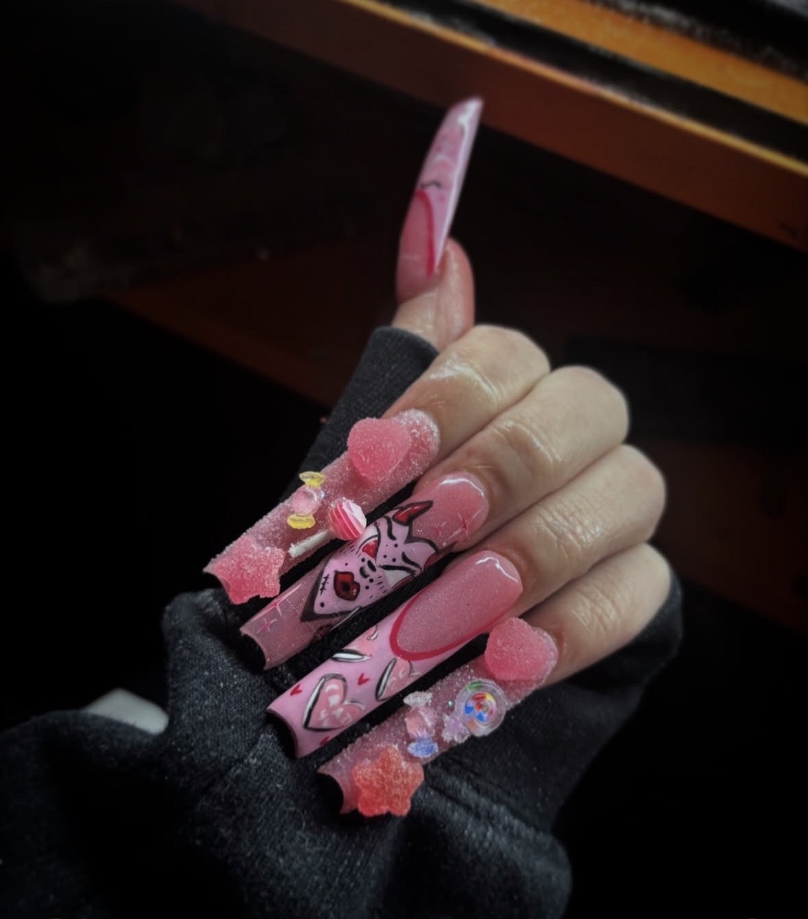 Hand with decorated long pink nails, wearing a black glove, holding a pink and black object, against a dark background.
