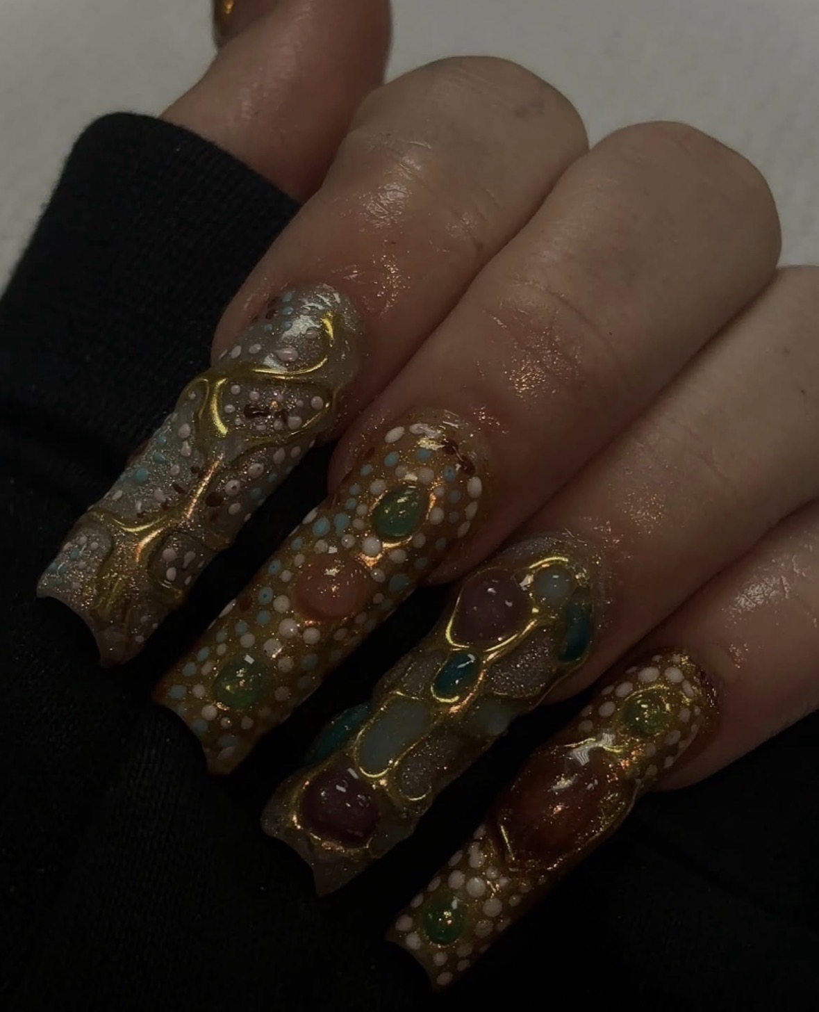 Close-up of a hand with long, decorated nails featuring intricate gold, green, and purple designs, and small embellishments.