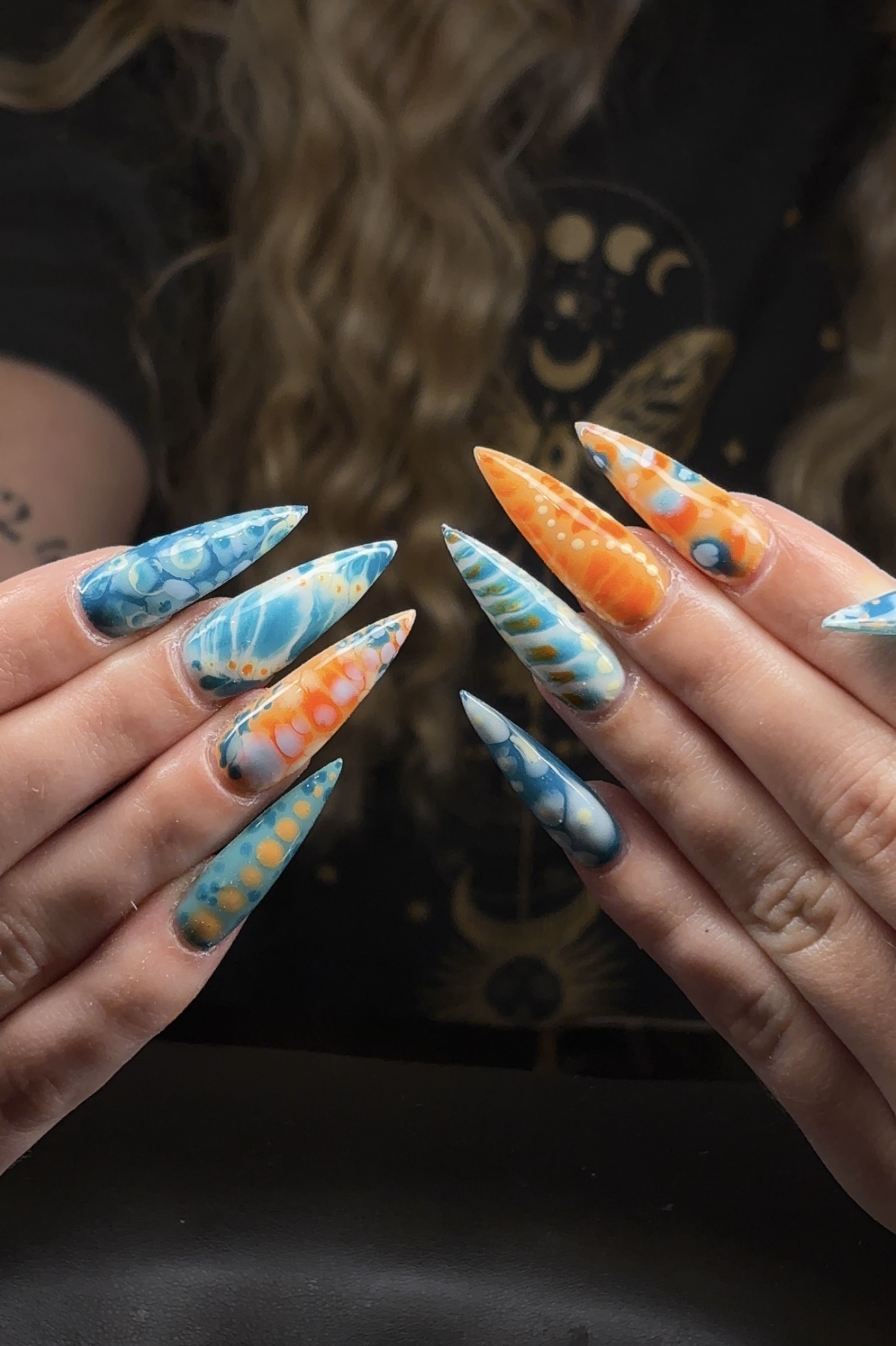 Person with long, wavy hair showing colorful, patterned, long, pointed nails in blue, orange, and white. Background is dark.