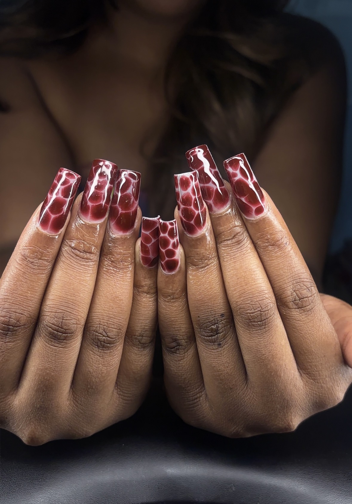 Person with long, wavy hair showing hands with red and white patterned nail art, fingers close together, background blurred.