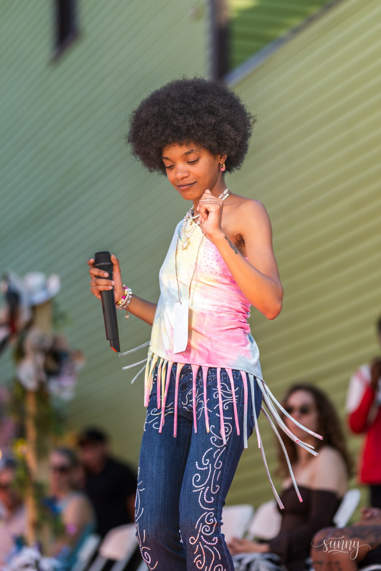 Young girl with curly hair holding a microphone, wearing a pink top and patterned pants, standing outdoors with people in the background.