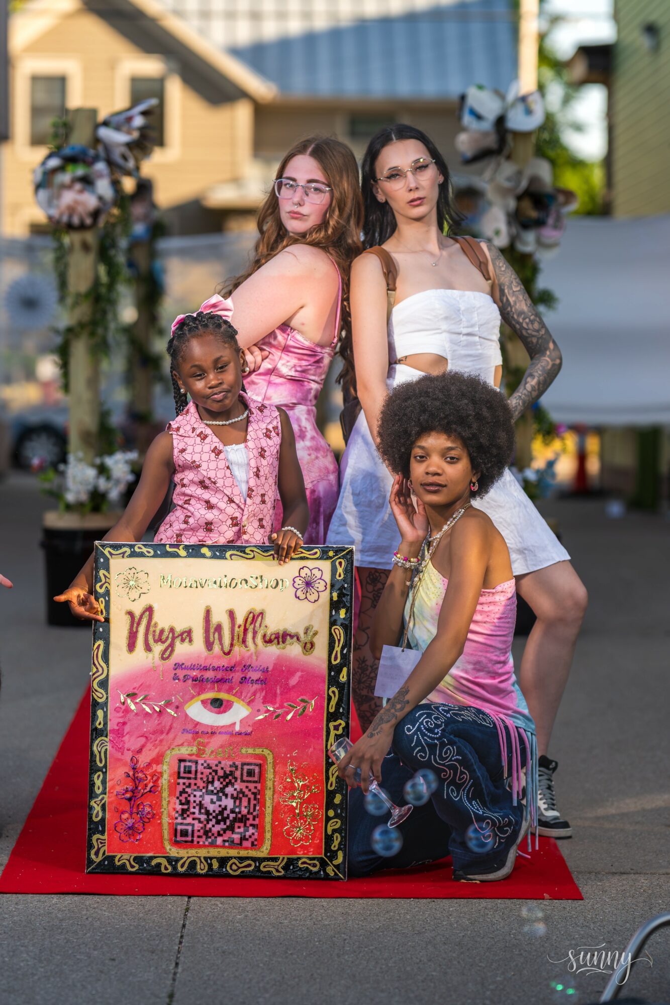 Four girls pose outdoors behind a decorated sign with a QR code, flowers, and eye illustration, on a red carpet.