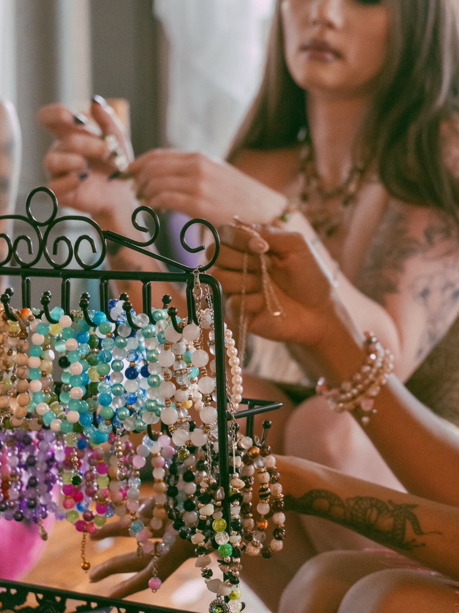 Woman with long hair and tattoos arranging colorful jewelry on a display stand, wearing bracelets and a necklace.