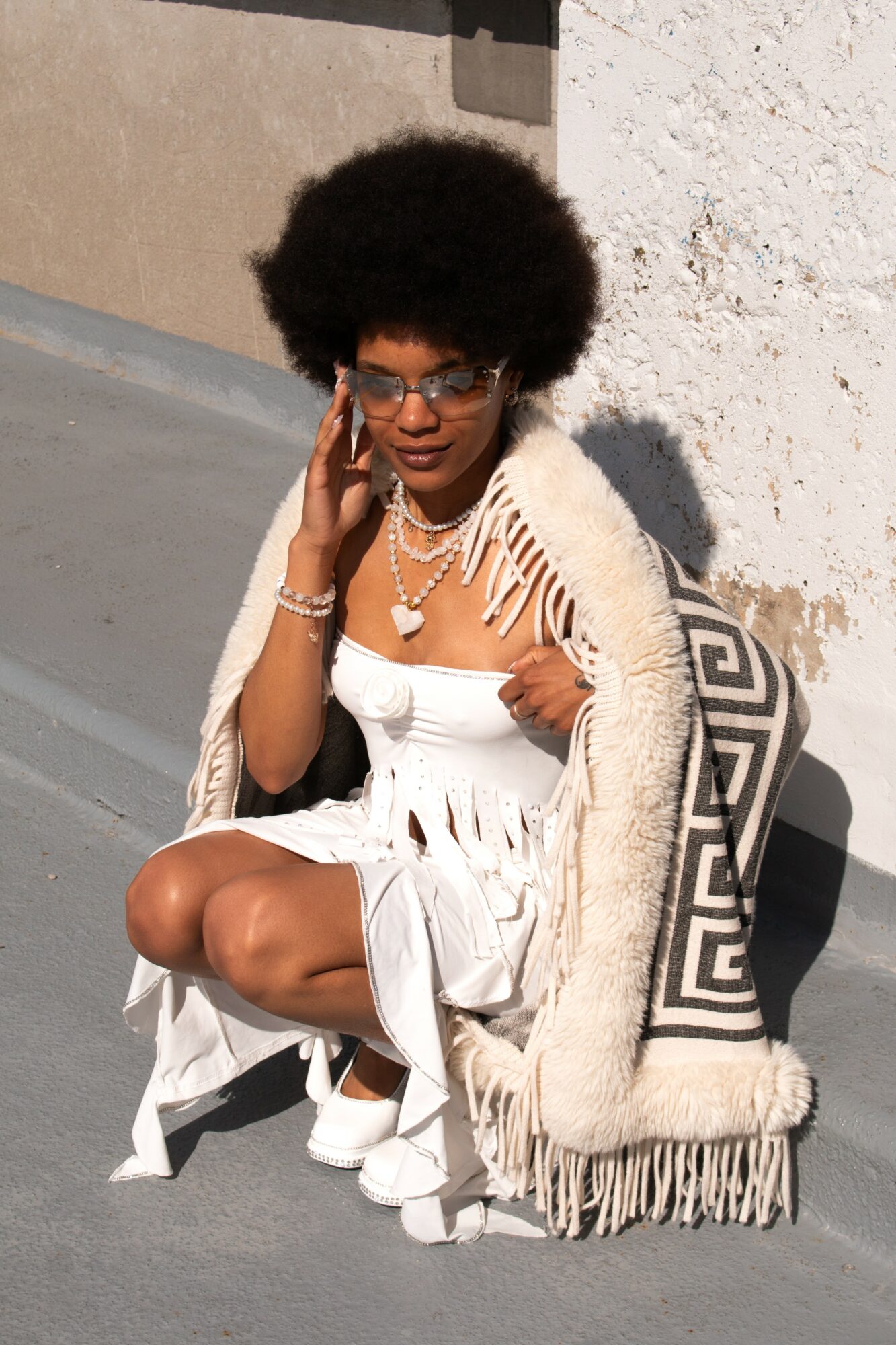 Woman with large afro hairstyle, glasses, and jewelry, sitting on the ground with a patterned blanket, outdoors on a sunny day.