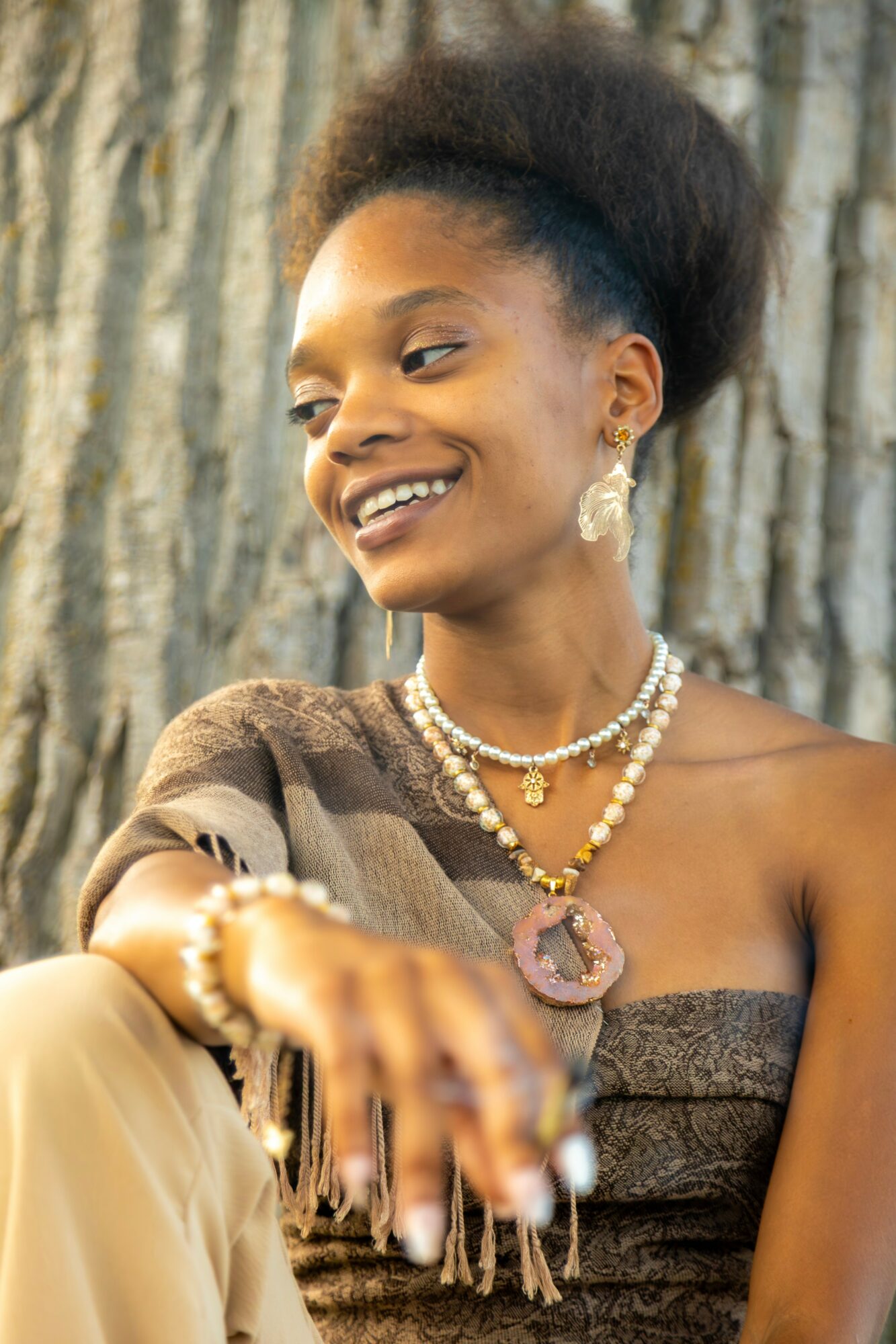 Smiling woman with natural hair, wearing jewelry and a strapless dress, outdoors with tree bark background.