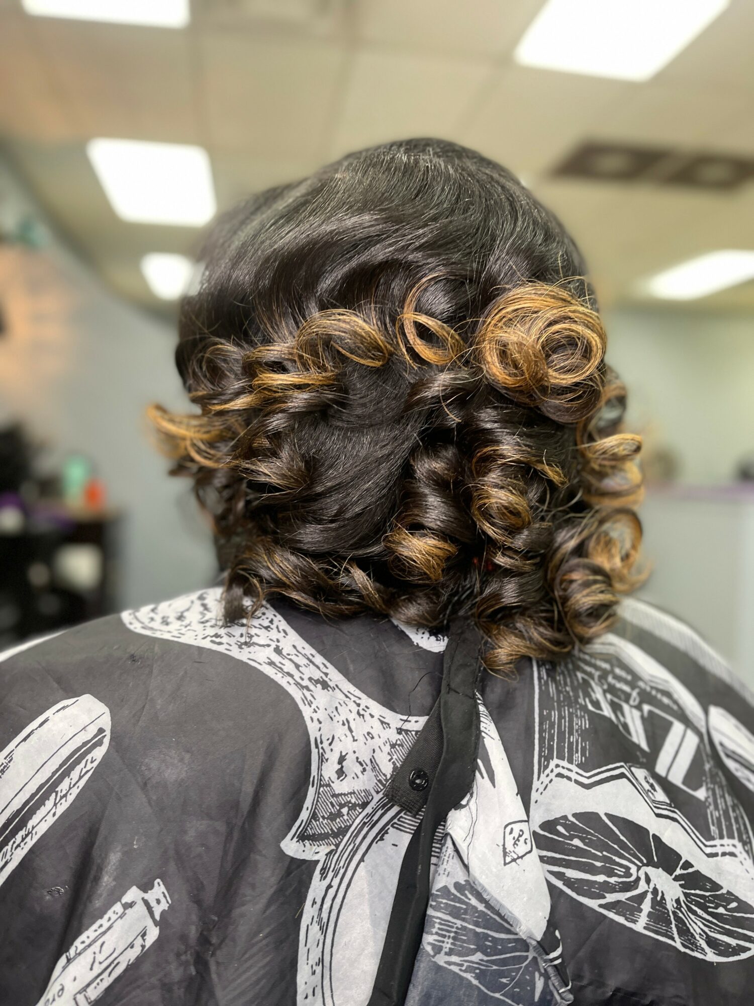 Back view of a woman with styled, curly hair featuring dark and light brown highlights, in a salon setting.