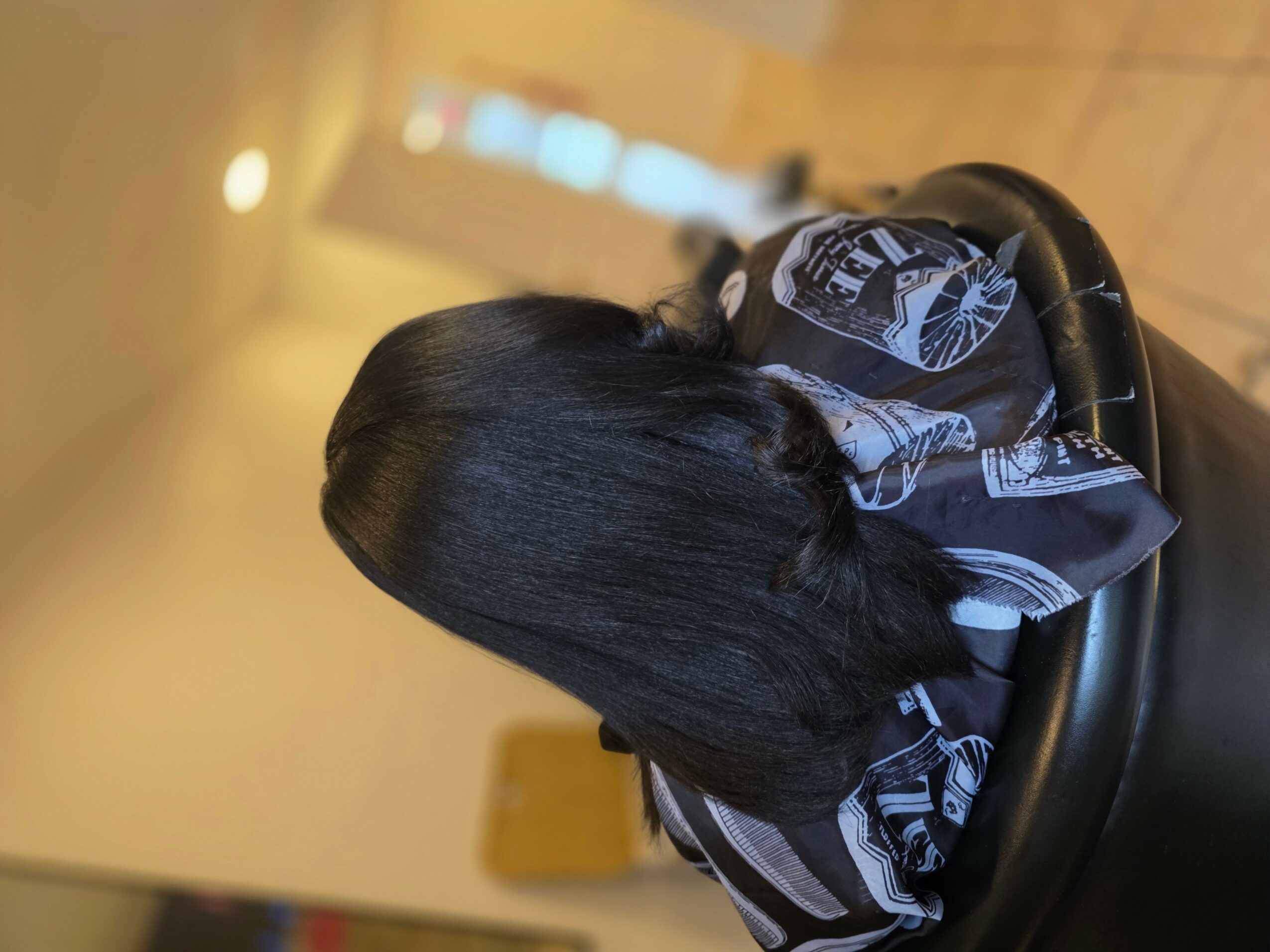 Person with long, straight black hair sitting in a chair, facing away, indoors with warm lighting