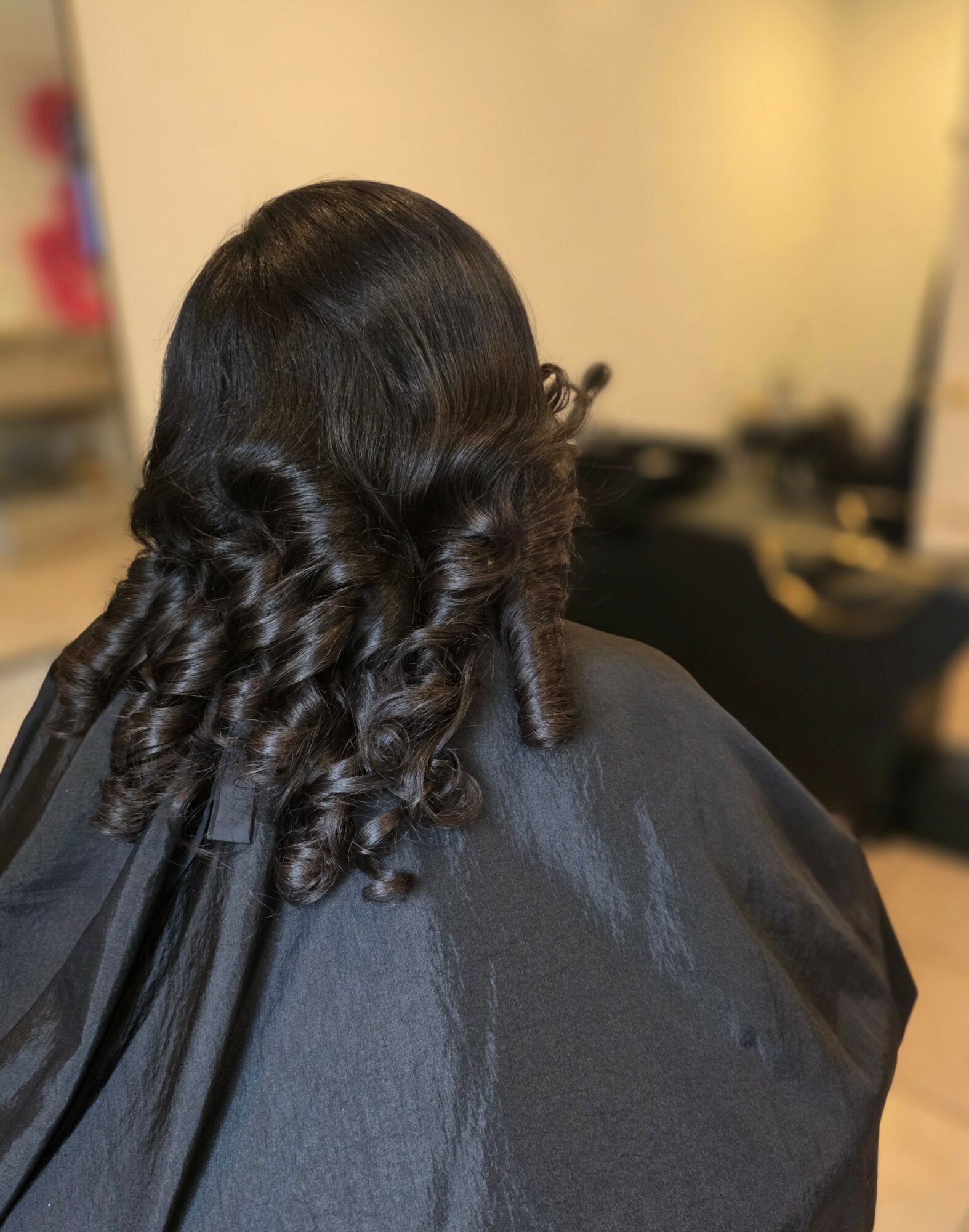 Back of a person's head with dark, curly hair, wearing a black cape, in a room with beige walls.