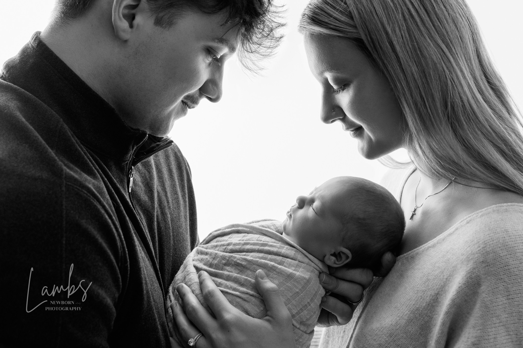 A man and woman look at a sleeping baby wrapped in a blanket, held close to their faces.